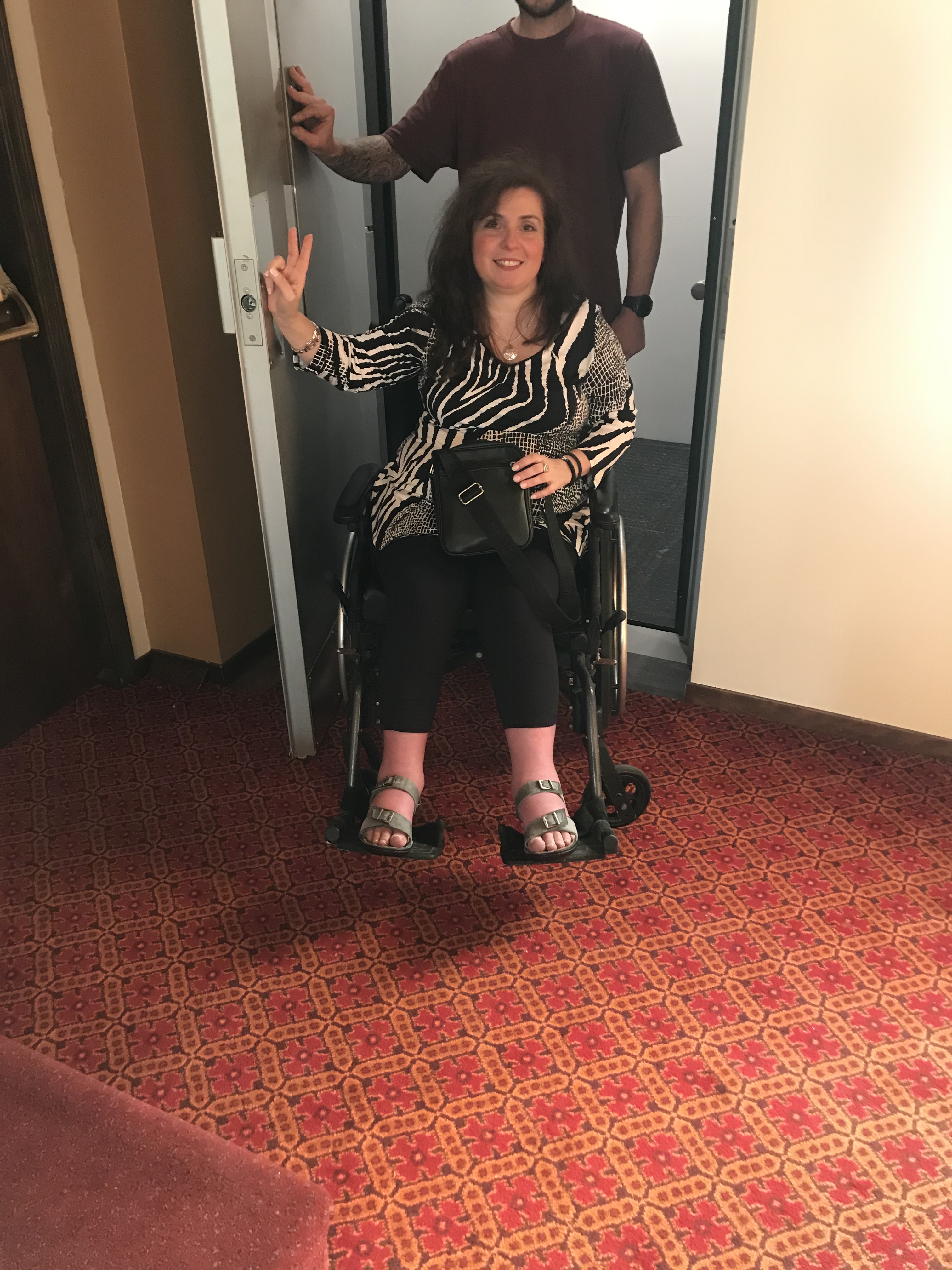 Person with long dark hair using a wheelchair entering through a doorway and giving the peace symbol with their fingers. 